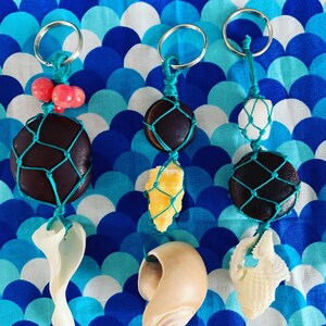 May include: Three handmade keychains with natural elements. Each keychain features a dark brown stone or a white stone encased in a teal net, paired with a shell or a cluster of pink beads. The keychains are suspended from silver key rings.