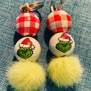 May include: Two keychains with red and white checkered spheres, white spheres with a Grinch face wearing a Santa hat, and yellow fluffy pom-poms. The keychains have silver rings.