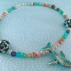 May include: A colorful beaded necklace featuring a silver whale pendant. The necklace has a variety of beads in shades of pink, blue, green, and brown, with two large black and white patterned beads. The necklace is a unique piece of jewelry.