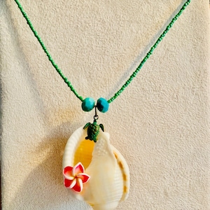 May include: A handcrafted necklace featuring a large, white seashell pendant adorned with a red and pink flower and a small green turtle charm. The necklace has a green beaded chain and turquoise beads.