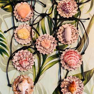 May include: A collection of seashell necklaces. Each necklace features a unique seashell in shades of pink, orange, and white, set on a decorative, lace-patterned backing. The shells are strung on black cords.