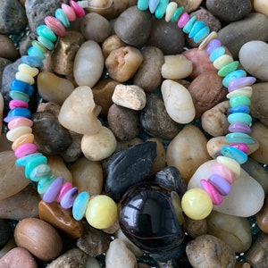May include: A colorful beaded bracelet featuring a variety of pastel-colored stones, including pink, blue, green, and yellow. The bracelet also includes a large, dark, rounded stone and two yellow beads. The bracelet is displayed on a bed of smooth, multicolored stones.