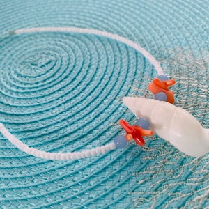 May include: White beaded necklace with a large white seashell pendant, coral-colored accents, and small blue beads. The necklace is displayed on a light blue textured surface, creating a coastal theme.
