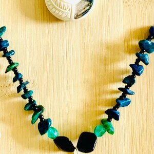 May include: Two necklaces featuring white carved pendants with spiral designs and iridescent shell inlays. The top necklace has light blue and green beads on a white cord. The bottom necklace features dark blue and green stone chips on a black cord.