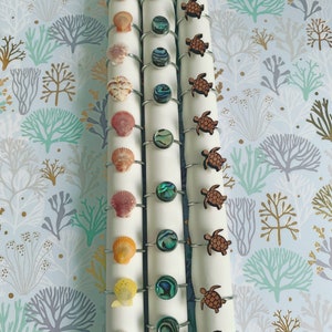 May include: A collection of napkin rings with ocean-themed designs. The rings feature seashells, abalone shells, and wooden sea turtles. The napkin rings are silver-toned and are displayed on white napkins against a blue and white patterned background.