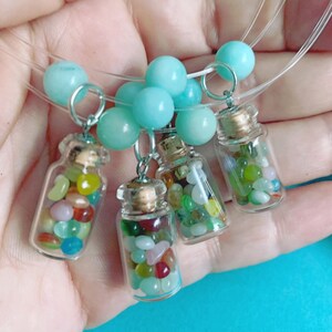 May include: Four miniature glass bottle pendants filled with colorful stones, each suspended from a strand of clear cord and light blue beads. The bottles have cork stoppers and silver-tone metal loops.