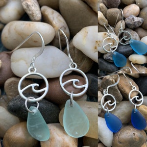 May include: Silver dangle earrings featuring a wave design within a circle, paired with teardrop-shaped sea glass in shades of blue and teal. The earrings have a hook closure and are displayed against a backdrop of smooth stones.