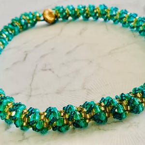 May include: A handcrafted necklace featuring a spiral design. The necklace is made with teal and gold beads, creating a textured, eye-catching pattern. The clasp is a small, round, gold-colored bead.