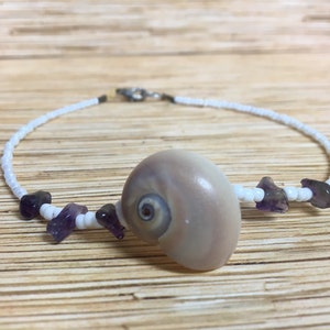 May include: A delicate white beaded bracelet featuring a large, light-colored seashell as a centerpiece. Accented with small, irregularly shaped purple gemstones. The bracelet is secured with a silver-colored clasp.