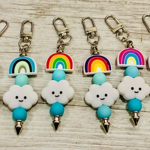 May include: Six keychains with rainbow and cloud charms. Each keychain has a silver clasp, a rainbow-shaped charm, a light blue bead, a white cloud charm with a smiling face, another light blue bead, and a silver cone-shaped charm.