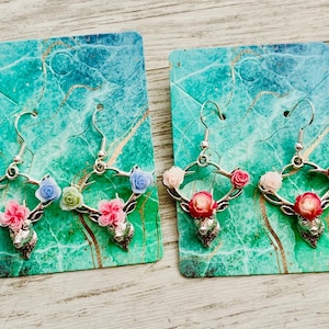 May include: Four pairs of floral antler earrings displayed on teal and green marbled cards. The earrings feature silver-toned antlers adorned with colorful miniature roses and a small skull pendant. Each pair has a unique color scheme.