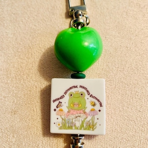 May include: A keychain with a bright green heart-shaped bead and a square charm. The charm features a cartoon frog wearing a crown, surrounded by mushrooms and flowers, with the text "Naturally Introverted, Selectively Extroverted."