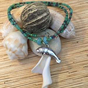 May include: A beaded necklace with a silver manatee pendant, featuring turquoise, green, and brown beads. The necklace is displayed with seashells and a textured stone on a wooden surface. The pendant is approximately 1 inch long.