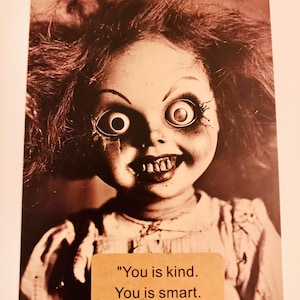 May include: Sepia-toned image of a vintage doll with wild, messy hair and a sinister grin. The doll has large, wide eyes and a distressed appearance. A small, rectangular sticker below the doll reads: "You is kind. You is smart. You is important."