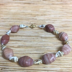 May include: A beaded bracelet featuring alternating pinkish-brown oval stones and small seashells. The bracelet is strung on a thin, silver-colored wire and has a gold-colored clasp. The stones have a striped pattern.