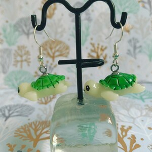 May include: A pair of turtle-shaped earrings. Each earring features a light green shell and a cream-colored body. The earrings hang from silver-colored fish hook ear wires. The earrings are displayed on a black metal stand.