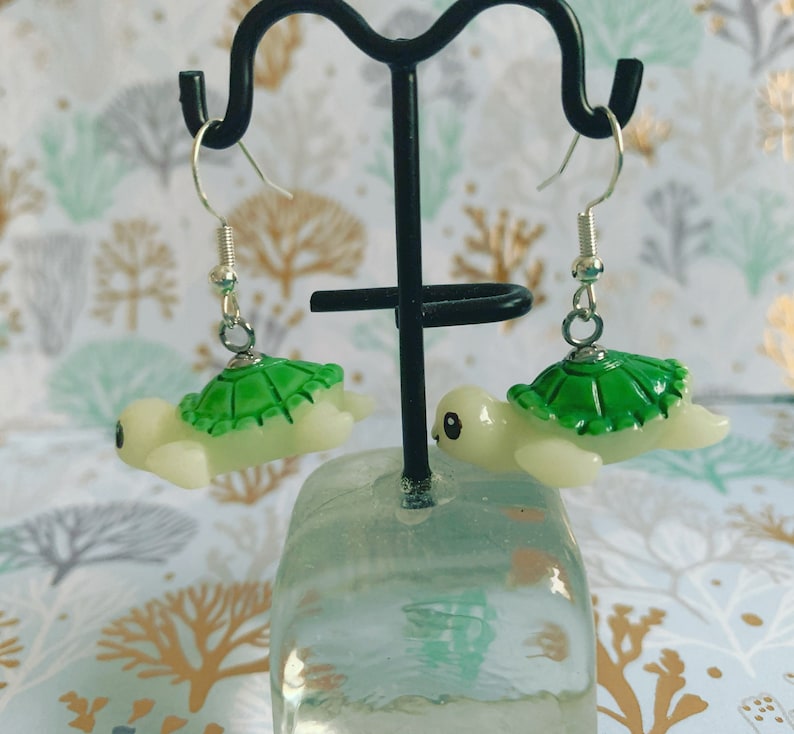 May include: A pair of turtle-shaped earrings. Each earring features a light green shell and a cream-colored body. The earrings hang from silver-colored fish hook ear wires. The earrings are displayed on a black metal stand.