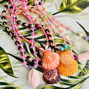 May include: Two layered necklaces featuring strands of small, translucent, multi-colored flower-shaped beads in shades of pink, purple, orange, and dark red. Each necklace is accented with large, natural seashells in peach, orange, and brown tones.