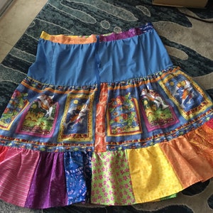 May include: A colorful patchwork skirt with a blue waistband and a tiered design. The skirt features a variety of patterned fabrics, including a blue fabric with framed illustrations, and a rainbow of solid and patterned panels in the lower tier.
