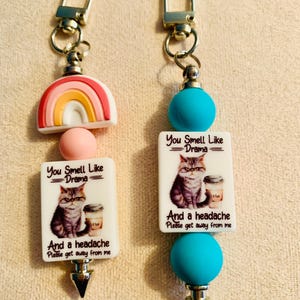 May include: Two keychains with silver-tone clasps. One features a rainbow charm, a pink bead, and a white rectangle with a cat graphic and text: "You Smell Like Drama And a headache Please get away from me." The other has a blue bead.