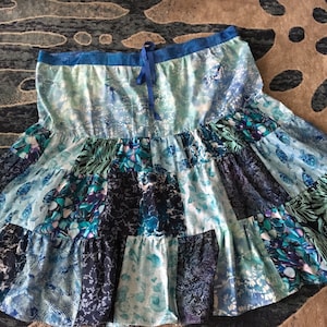 May include: A patchwork skirt in shades of blue, teal, and white. The skirt features a tiered design with various floral and abstract patterns. A blue ribbon is tied at the waist. The skirt is made of a lightweight fabric.