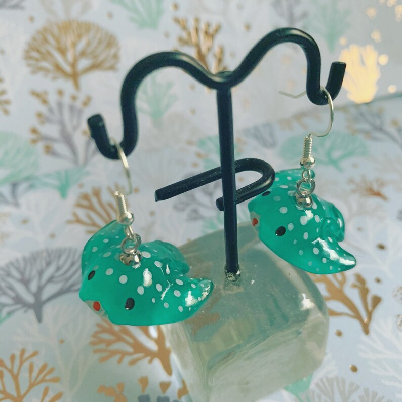 May include: A pair of teal stingray earrings with white spots and red accents. The earrings are displayed on a black metal stand with silver hooks. The background features a patterned design with gold and teal elements.