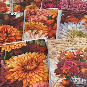 May include: A collection of greeting cards featuring vibrant floral designs. The cards showcase close-up images of chrysanthemums in shades of orange, yellow, pink, and white, with some cards displaying a basket of autumn flowers and text.