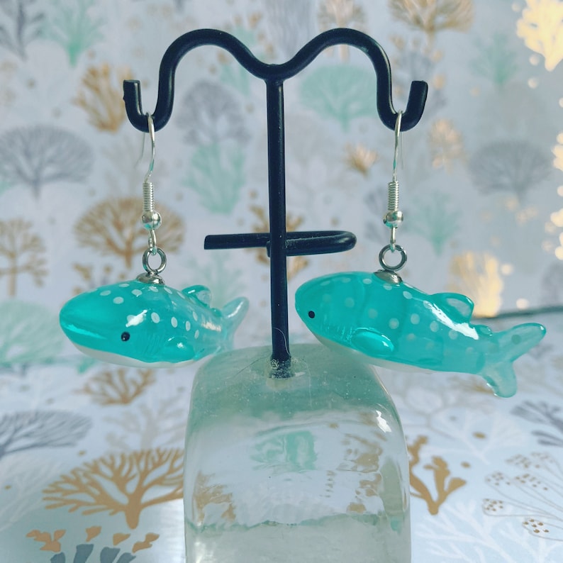 May include: A pair of turquoise whale shark earrings. Each earring features a detailed, miniature whale shark with white spots and a small black eye. The earrings hang from silver-colored fish hook ear wires.