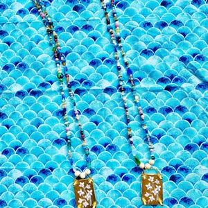 May include: Two beaded necklaces with rectangular pendants. The pendants are gold-colored with a floral design in white. The necklaces have a variety of colorful beads. The background is a blue fabric with a scale pattern.
