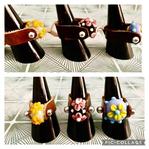 May include: Handmade leather rings with colorful glass bead designs. The rings feature brown leather bands and are adorned with yellow, pink, and blue floral-shaped beads. Each ring is displayed on a black cone-shaped stand.