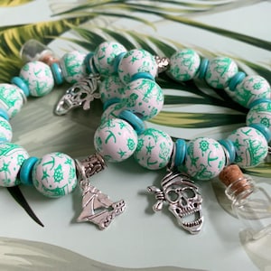 May include: A beaded bracelet with a pirate theme. The bracelet features white beads with green nautical designs, turquoise spacers, and silver charms including a skull, ship, and anchor. A small glass bottle with a cork is also included.