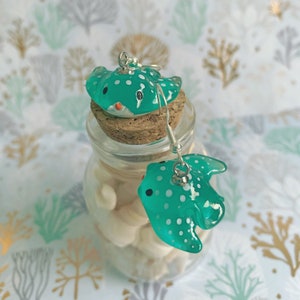 May include: A pair of handmade teal stingray earrings with white polka dots. The earrings are made of a translucent material and feature silver-colored ear wires. The stingrays have black eyes and orange accents. The earrings are displayed on a cork and glass jar.