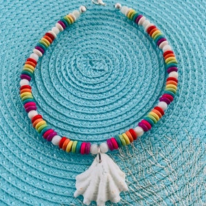 May include: A colorful necklace with a white seashell pendant. The necklace features a strand of rainbow-colored beads, including red, orange, yellow, green, blue, and purple, interspersed with white beads. The pendant is a textured, white seashell.