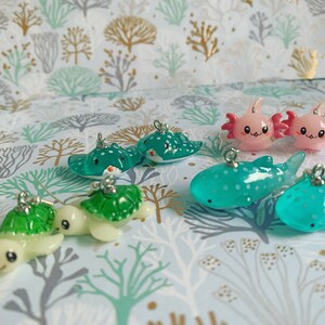 May include: A collection of miniature ocean-themed charms. The charms include sea turtles, stingrays, axolotls, and whale sharks in various shades of green, pink, and white. Each charm has a silver loop for attaching to jewelry or other crafts.