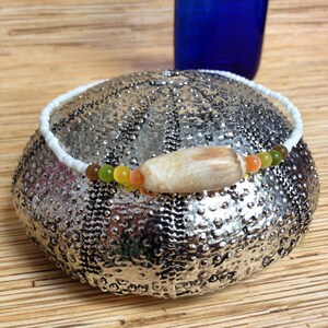 May include: A white beaded necklace with a central, oblong, beige shell pendant. The necklace features a strand of small, colorful beads in shades of green, orange, and brown. The necklace is displayed on a textured, silver-colored surface.