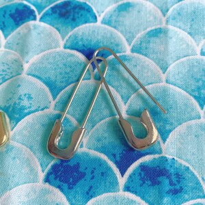 Puede incluir: Dos imperdibles plateados se muestran sobre una tela estampada azul y blanca. Los imperdibles est&aacute;n abiertos, mostrando su dise&ntilde;o curvo. La tela tiene un patr&oacute;n repetitivo de formas azules y blancas, que se asemejan a escamas.