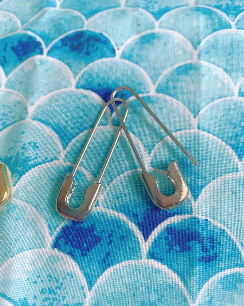 Puede incluir: Dos imperdibles plateados se muestran sobre una tela estampada azul y blanca. Los imperdibles est&aacute;n abiertos, mostrando su dise&ntilde;o curvo. La tela tiene un patr&oacute;n repetitivo de formas azules y blancas, que se asemejan a escamas.