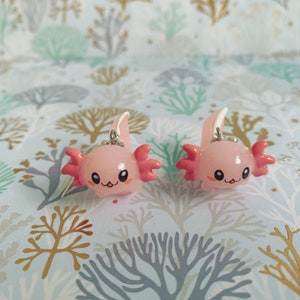 May include: Two pink axolotl charms with silver-colored metal loops for hanging. Each charm has a smiling face, pink gills, and a white and pink fin. The axolotls are set against a patterned background.
