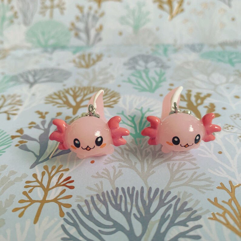 May include: Two pink axolotl charms with silver-colored metal loops for hanging. Each charm has a smiling face, pink gills, and a white and pink fin. The axolotls are set against a patterned background.