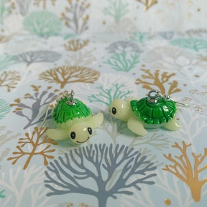 May include: Two pairs of turtle earrings. Each earring features a green turtle shell and a cream-colored body. The turtles have smiling faces and are suspended from silver-colored hooks. The earrings are set against a patterned background.