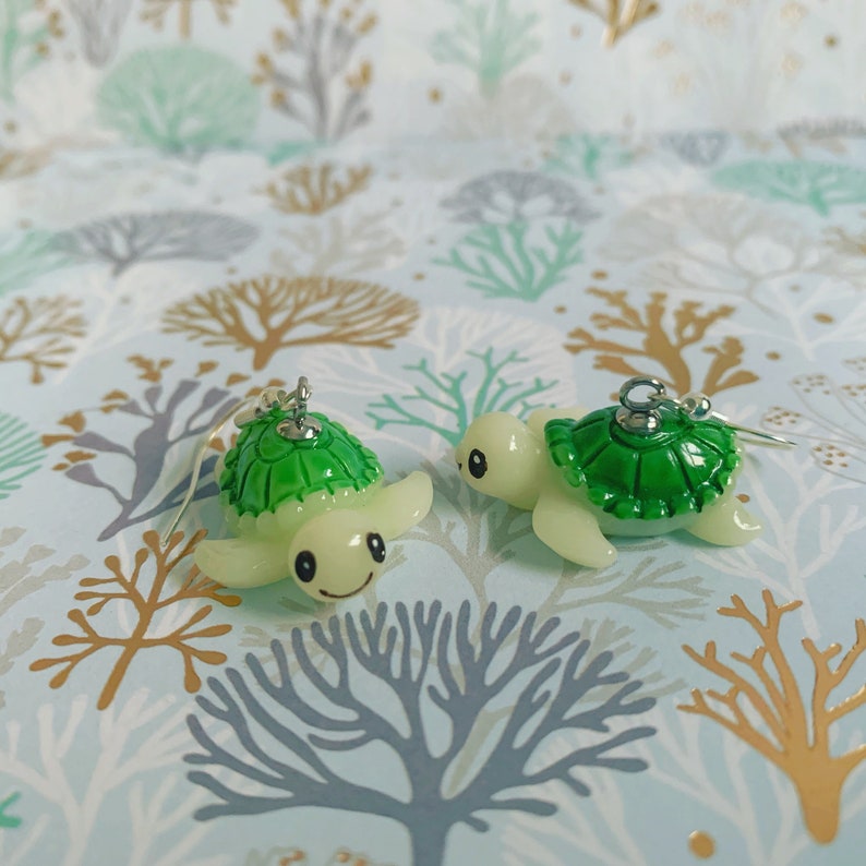 May include: Two pairs of turtle earrings. Each earring features a green turtle shell and a cream-colored body. The turtles have smiling faces and are suspended from silver-colored hooks. The earrings are set against a patterned background.
