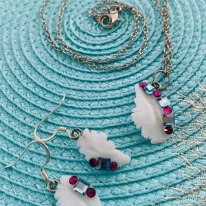 May include: A jewelry set featuring a white feather pendant necklace and matching earrings. The feathers are adorned with pink and blue gemstones, set on a silver chain and earring hooks. The set is displayed on a light blue woven surface.