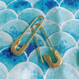 Puede incluir: Dos pendientes dorados con forma de imperdible se muestran sobre una tela estampada en azul y blanco. Los pendientes tienen forma de imperdibles, con un extremo curvado y un alfiler recto. La tela tiene un estampado de escamas.
