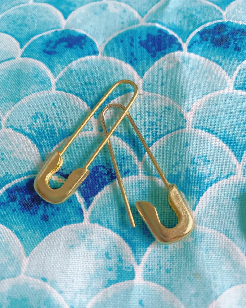 Puede incluir: Dos pendientes dorados con forma de imperdible se muestran sobre una tela estampada en azul y blanco. Los pendientes tienen forma de imperdibles, con un extremo curvado y un alfiler recto. La tela tiene un estampado de escamas.