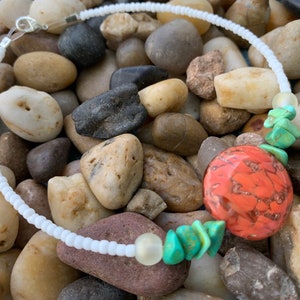 May include: A white beaded choker necklace with a large, orange and brown stone pendant. The necklace also features turquoise-colored stone accents and a silver clasp. The necklace is displayed on a bed of smooth, colorful stones.