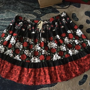 May include: A black skirt with a tiered design, featuring a repeating pattern of white skulls, red roses, and green leaves. The bottom tier is red with a rose pattern, and the skirt has a drawstring waist.