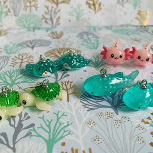 May include: A collection of miniature sea creature charms, including stingrays, turtles, axolotls, and whale sharks. The charms are in shades of green, blue, and pink, with silver-colored metal loops for attaching to jewelry or other crafts.