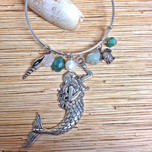 May include: A silver bangle bracelet with various charms, including a mermaid, a seashell, and several gemstones. The charms are suspended from a silver hoop. A large seashell rests on the wooden surface behind the bracelet.