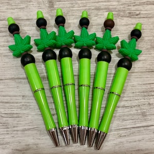 May include: A collection of six green pens with silver accents and black tips. Each pen is topped with a green cannabis leaf and a black and brown decorative element. The pens are arranged on a light wood-grain surface.