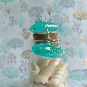 May include: A pair of turquoise whale shark earrings with white spots and silver hooks. The earrings are displayed on a cork and glass jar filled with seashells, against a patterned background.